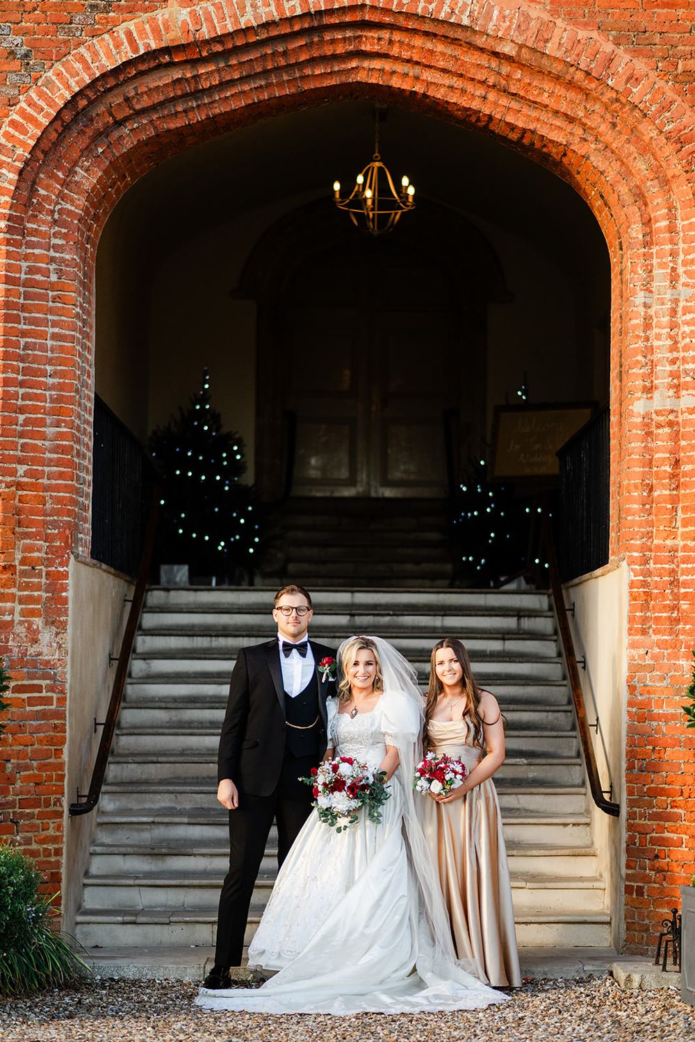 A wedding party poses under a grand brick archway with stone steps and elegant lighting in a historic venue.