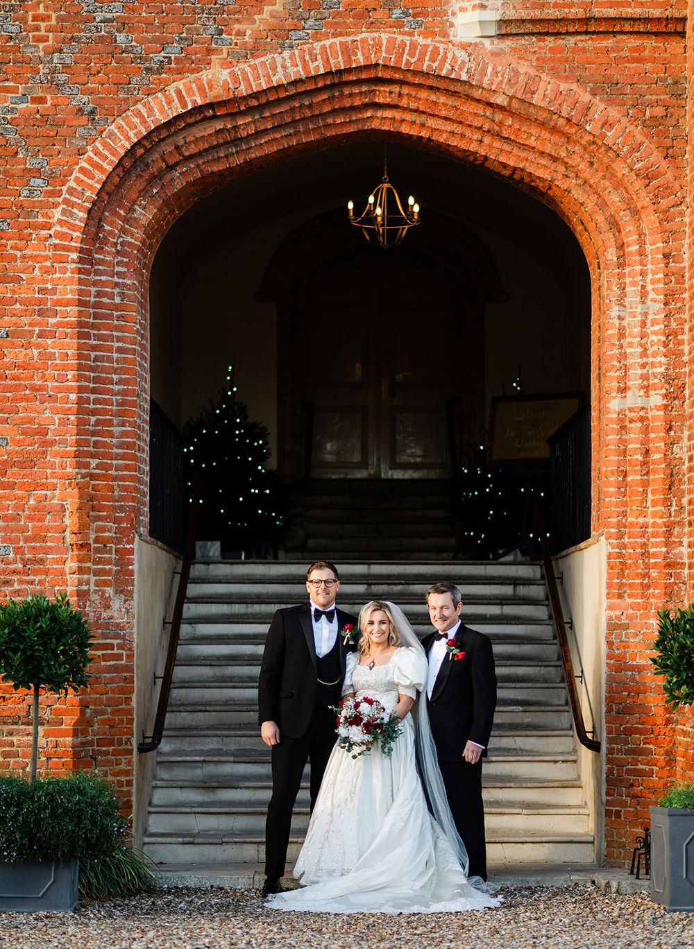 Elegant brick archway frames formal wedding photo with stone steps and decorative topiaries.