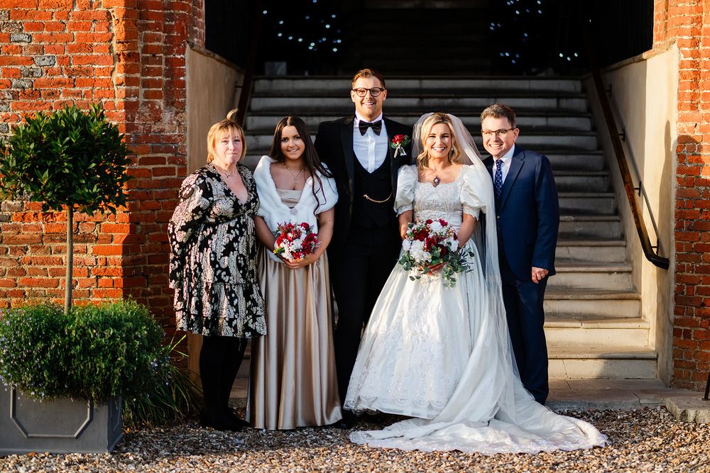 Wedding party poses in front of brick building with elegant formal attire and floral arrangements.