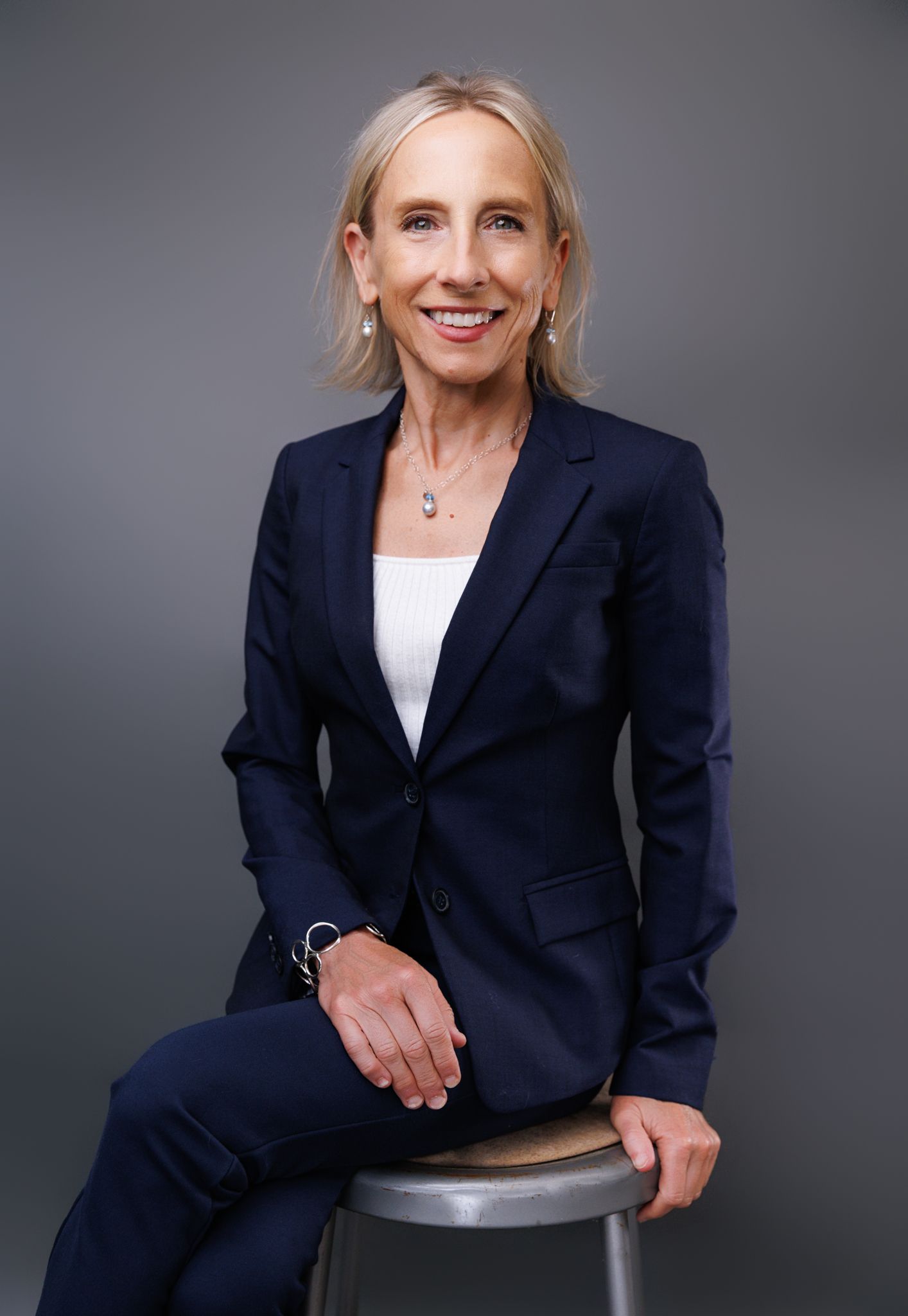 Professional woman in navy blue suit sits on stool smiling for corporate headshot portrait.