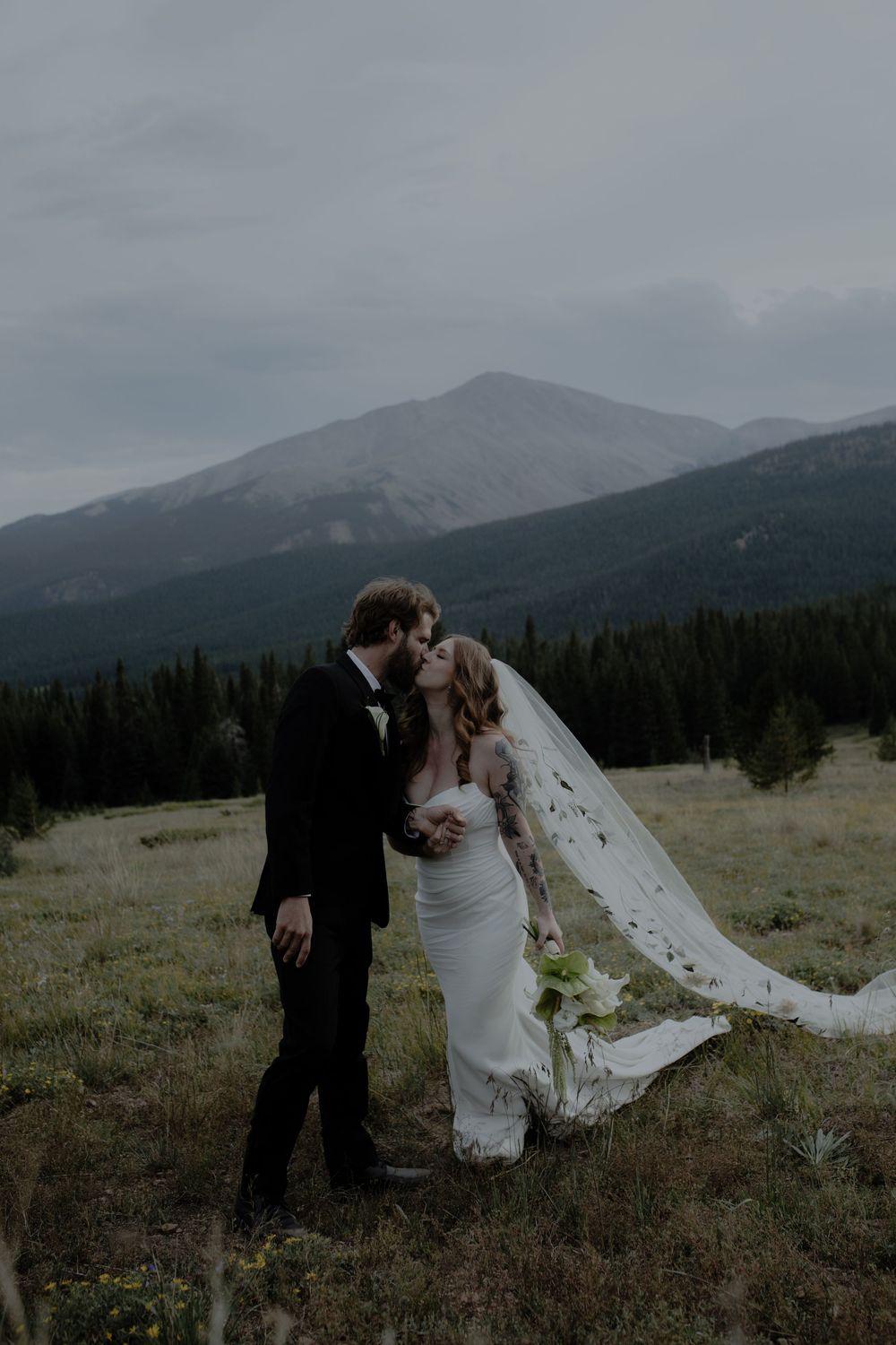 A couple shares an intimate moment in their wedding attire against a mountain landscape at dusk.