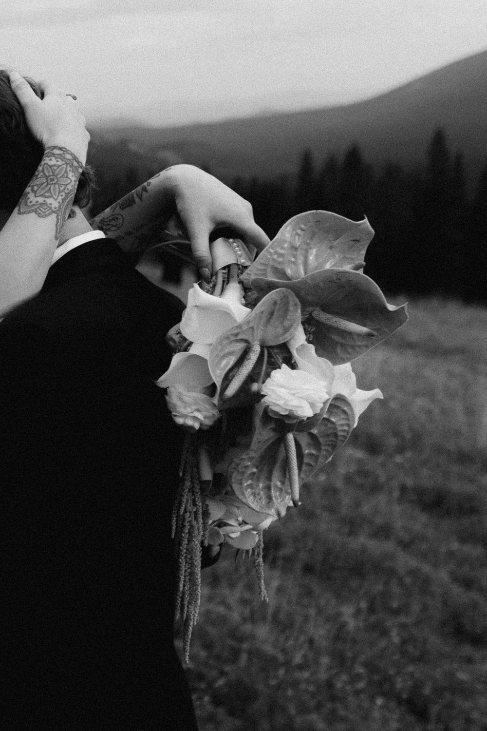 A black and white close-up of hands holding a delicate bouquet of roses against mountain scenery.
