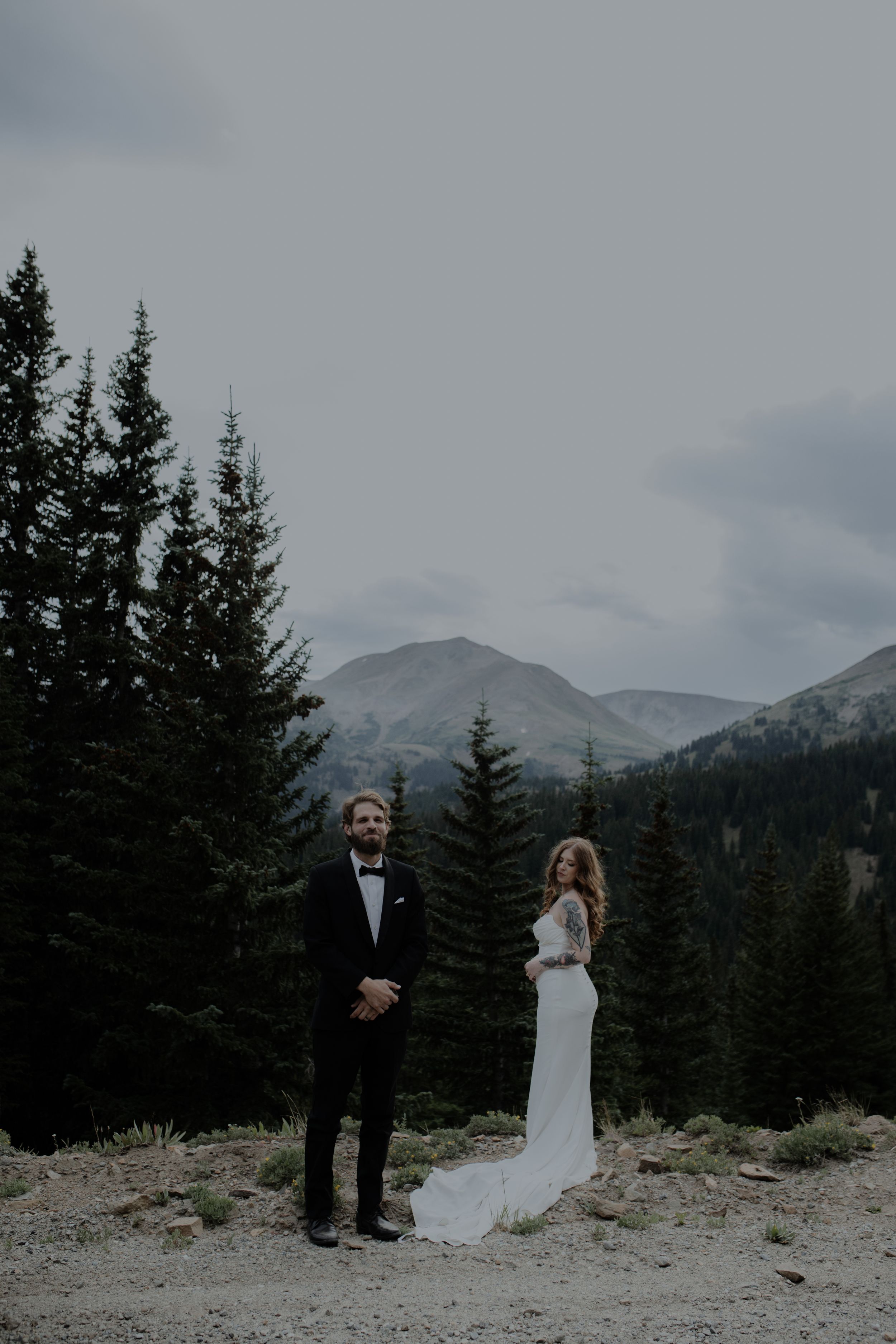 Wedding couple poses in front of evergreen trees and snow-capped mountains.