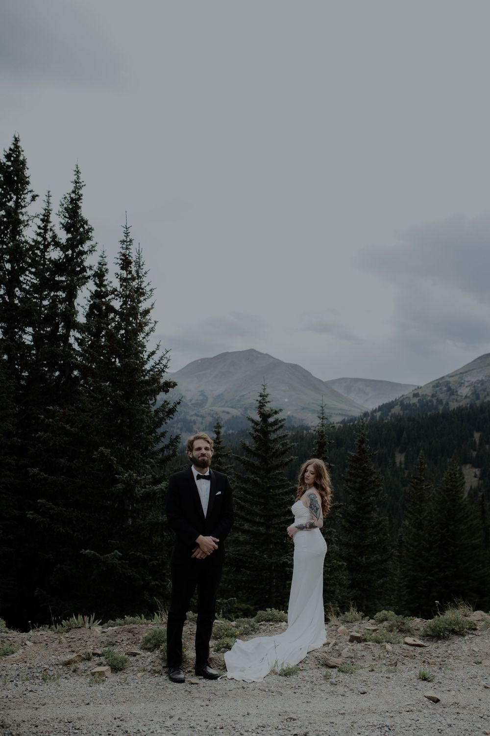 Wedding couple poses in front of evergreen trees and snow-capped mountains.