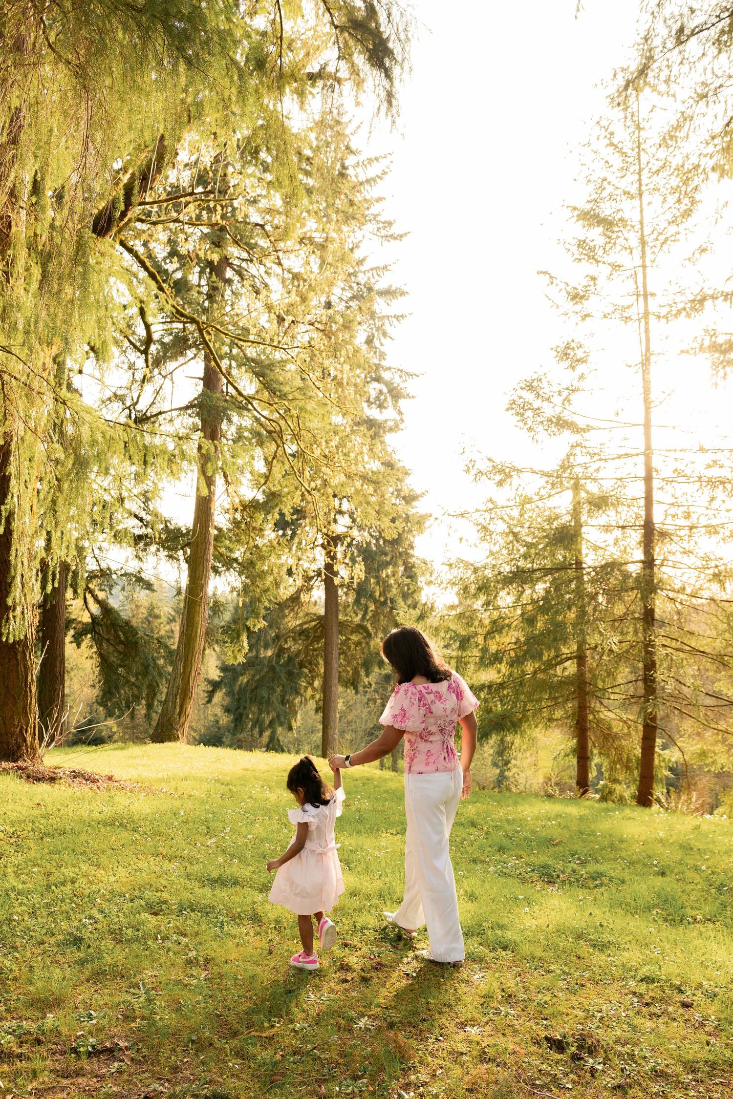 Two figures in matching pink outfits hold hands while walking on a grassy hill at sunset among tall evergreen trees.