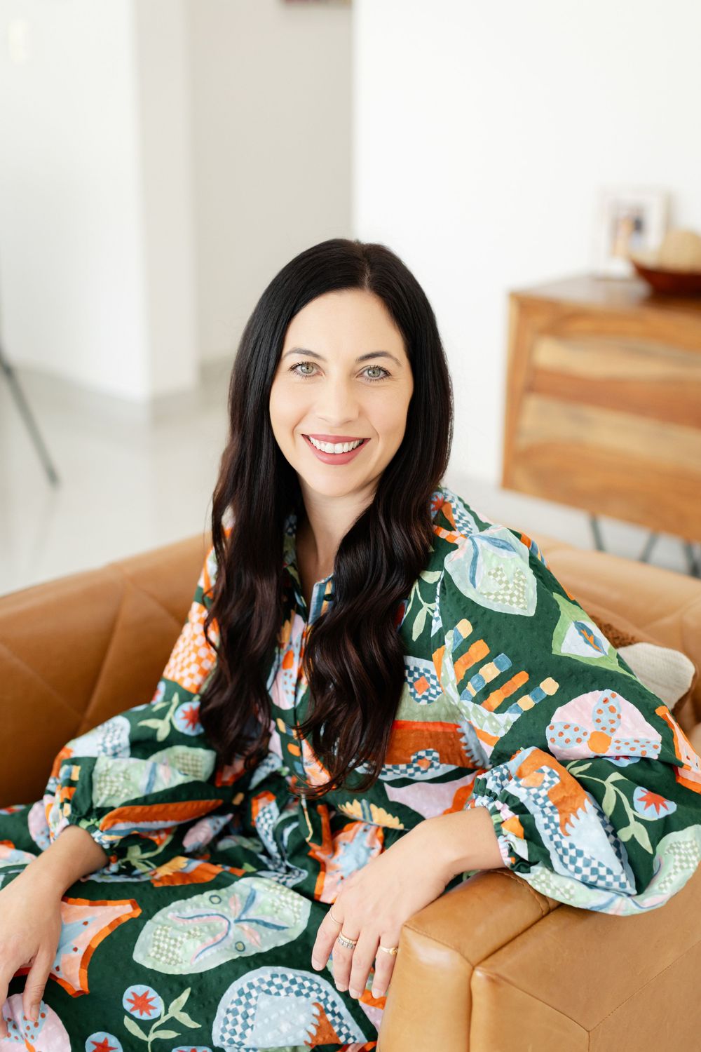 A woman in a colorful floral dress sits comfortably on a tan leather couch whilst having Branding Photos in Perth