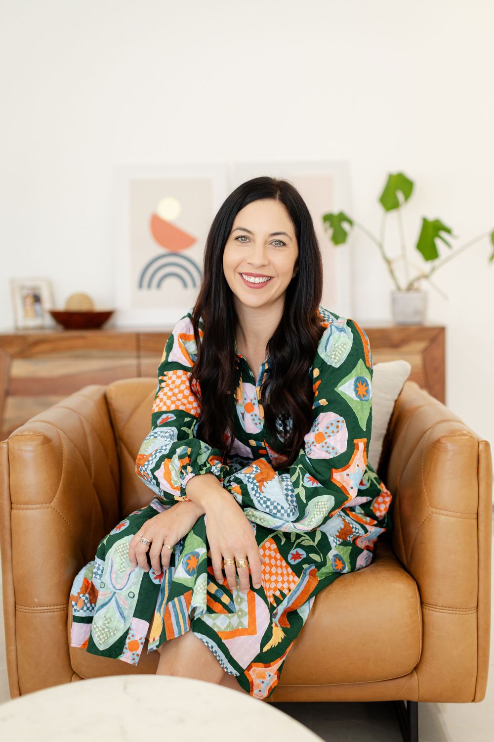 A woman in a colorful patterned dress sits smiling on a tan leather armchair in a Branding Photography Session
