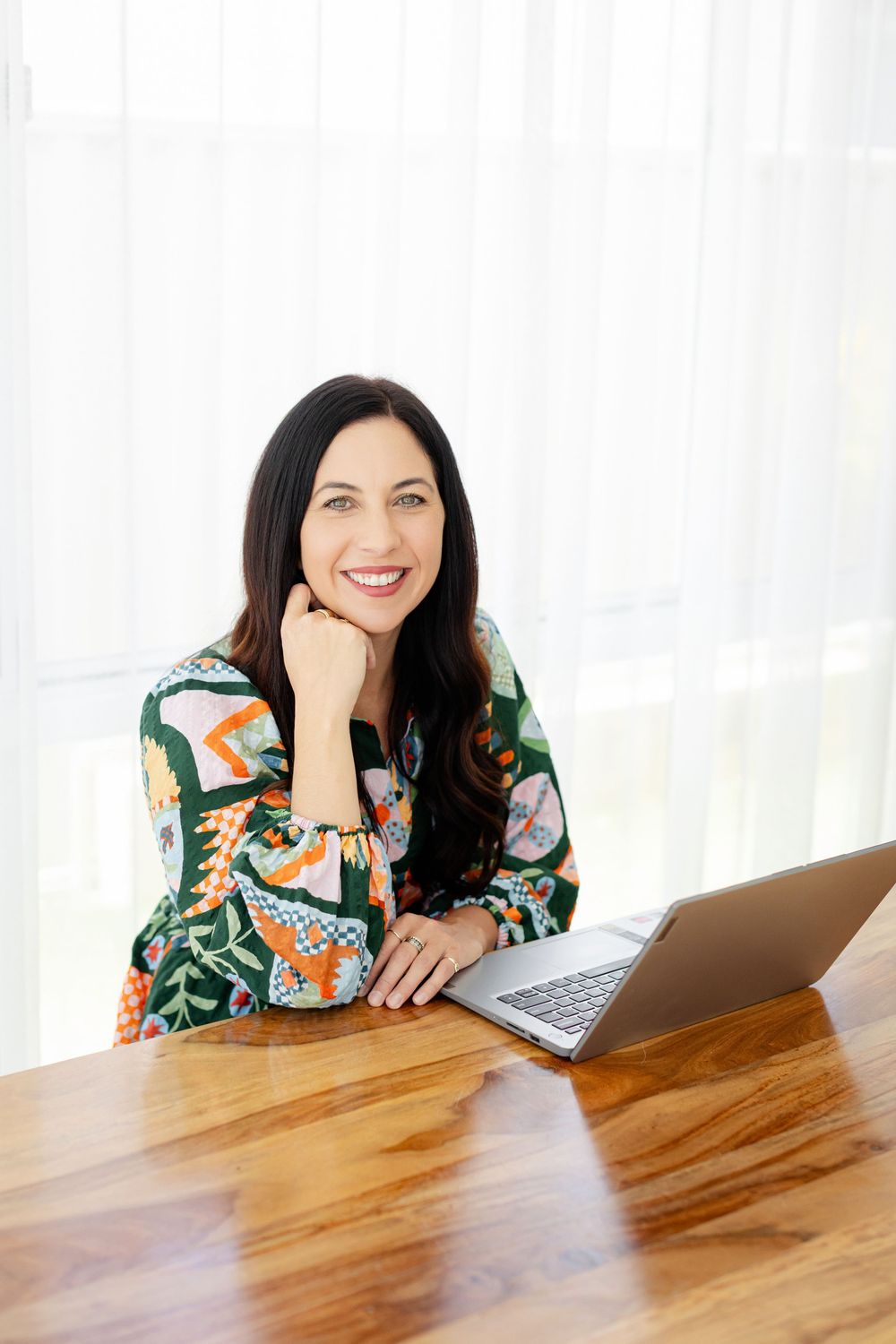 Woman in floral print top sits at wooden desk with laptop while having a branding shoot with Kirsten Graham Photography