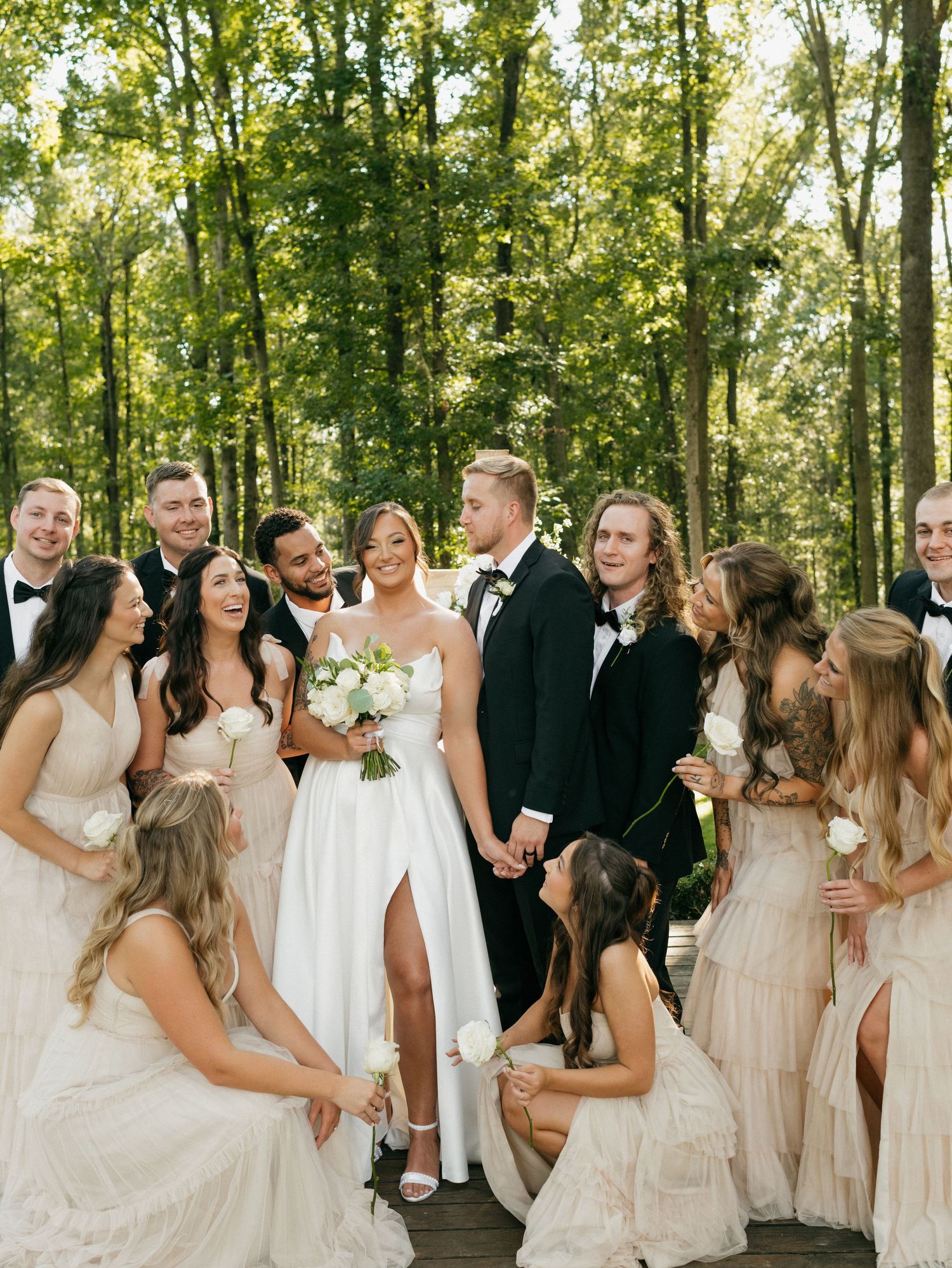 Wedding party in light colored dresses and dark suits laughs together in a wooded outdoor setting.