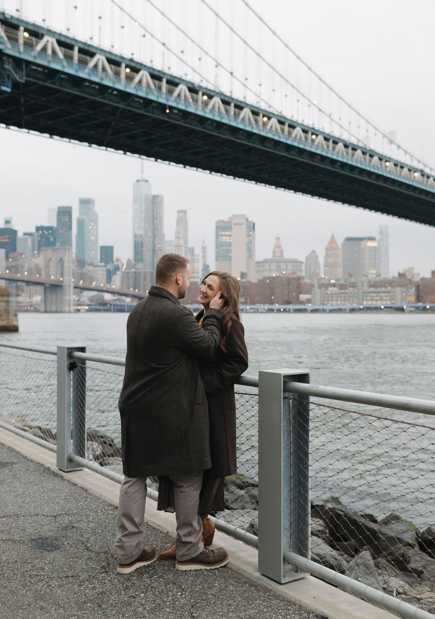 Romantic couple moment captured under Manhattan Bridge with city skyline in background.