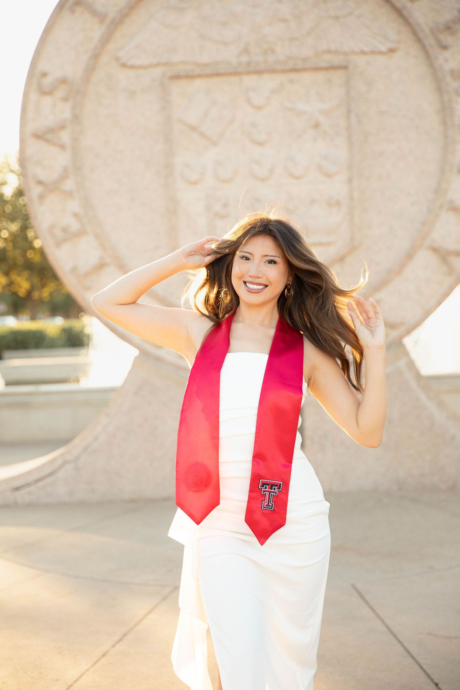 Graduate in white dress and red stole poses outdoors during sunset near ornate stone architecture.