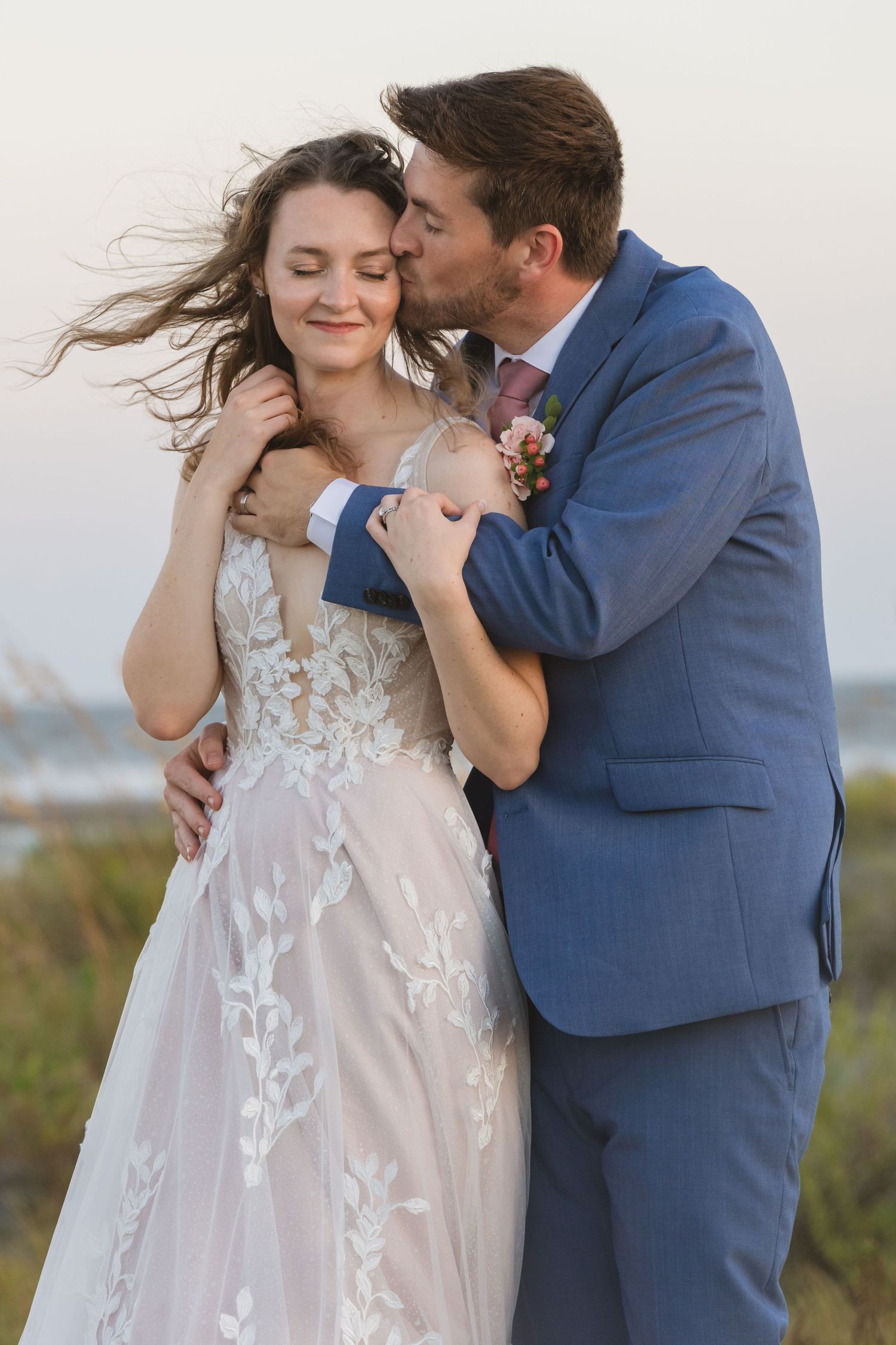 Romantic wedding couple embracing in blue suit and lace dress with wind-swept hair.