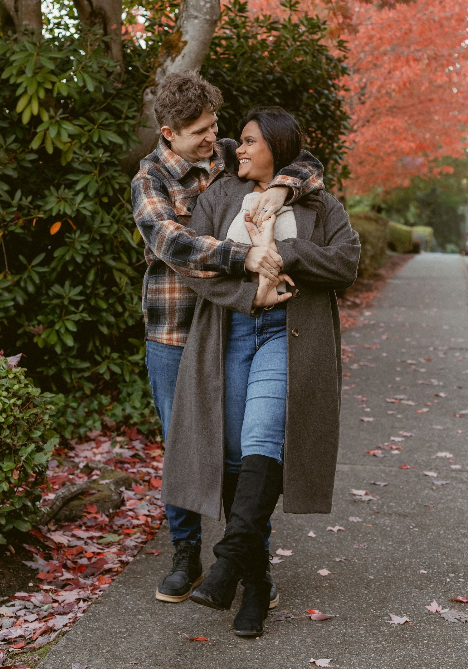 A couple embraces on a pathway surrounded by autumn foliage and fallen red leaves on the ground.