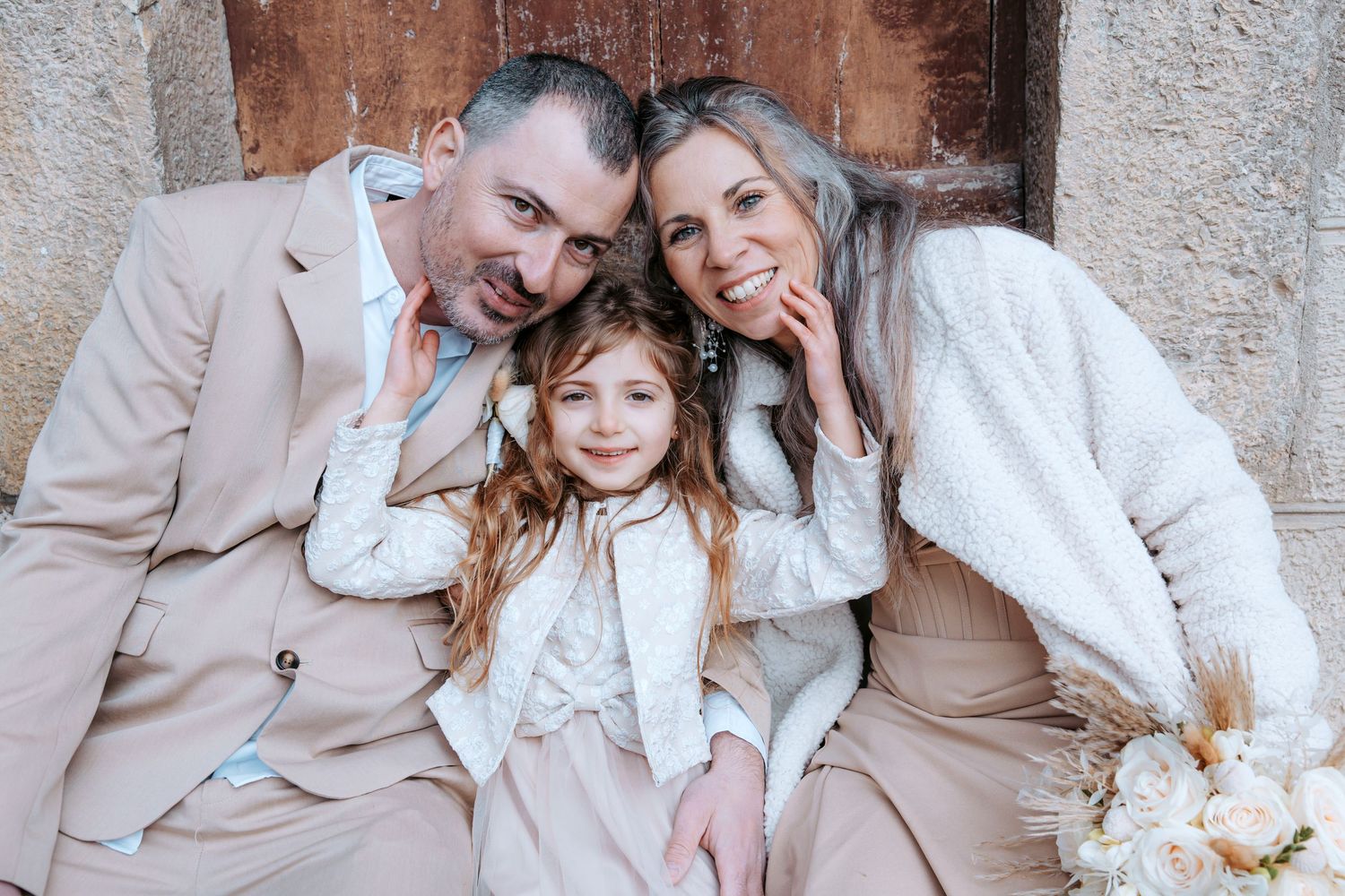 A family in neutral beige outfits poses together against a rustic stone wall with white floral bouquet.