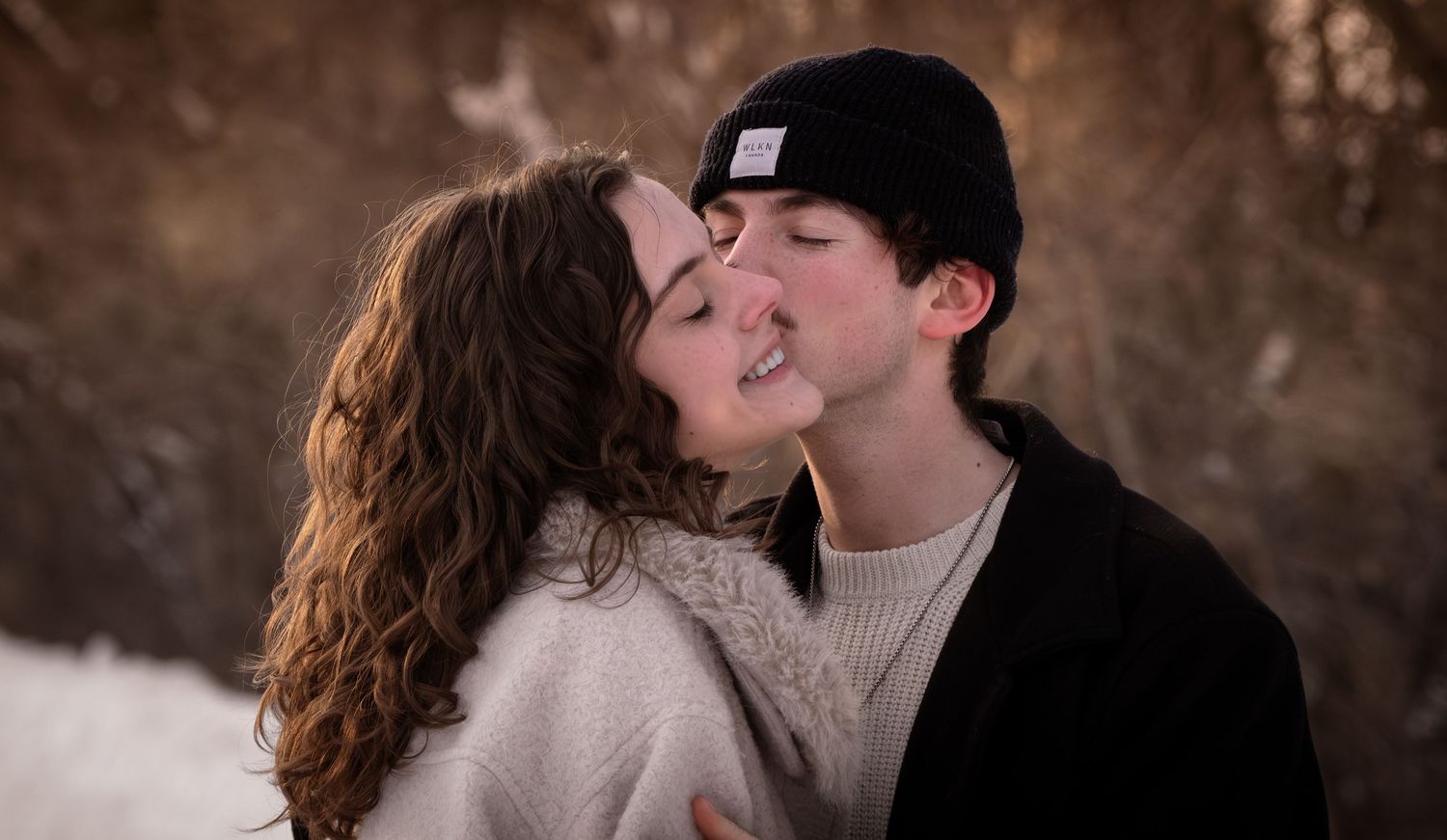Couple sitting back to back laughing in snowy winter setting wearing cozy sweaters.