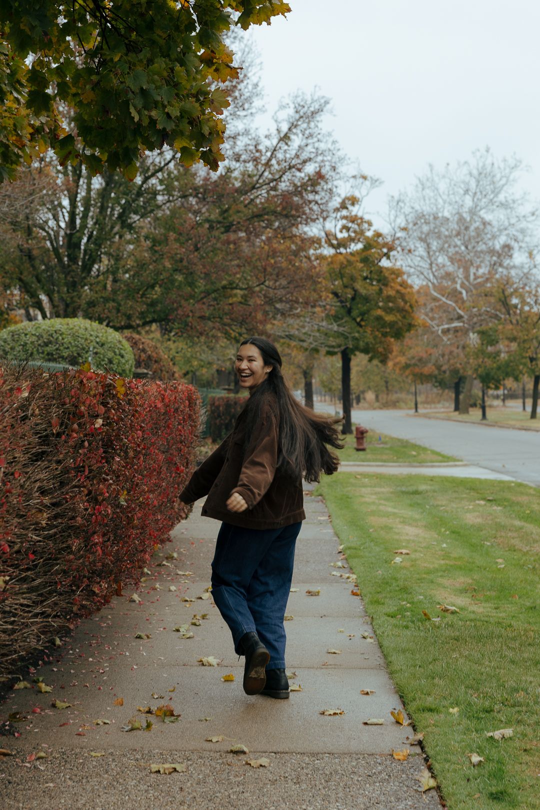 A person in dark clothing walks along an autumn sidewalk bordered by red bushes and trees with falling leaves.
