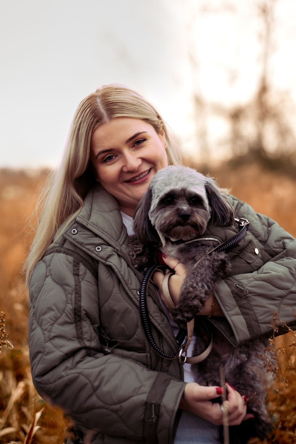 Person in olive green jacket cuddling with small grey dog outdoors.