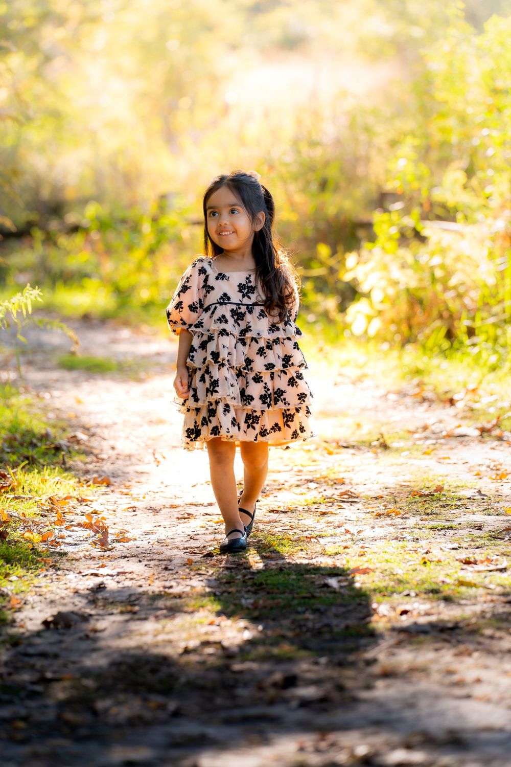 A young child in a floral dress runs playfully along a sunlit dirt path in a natural outdoor setting.
