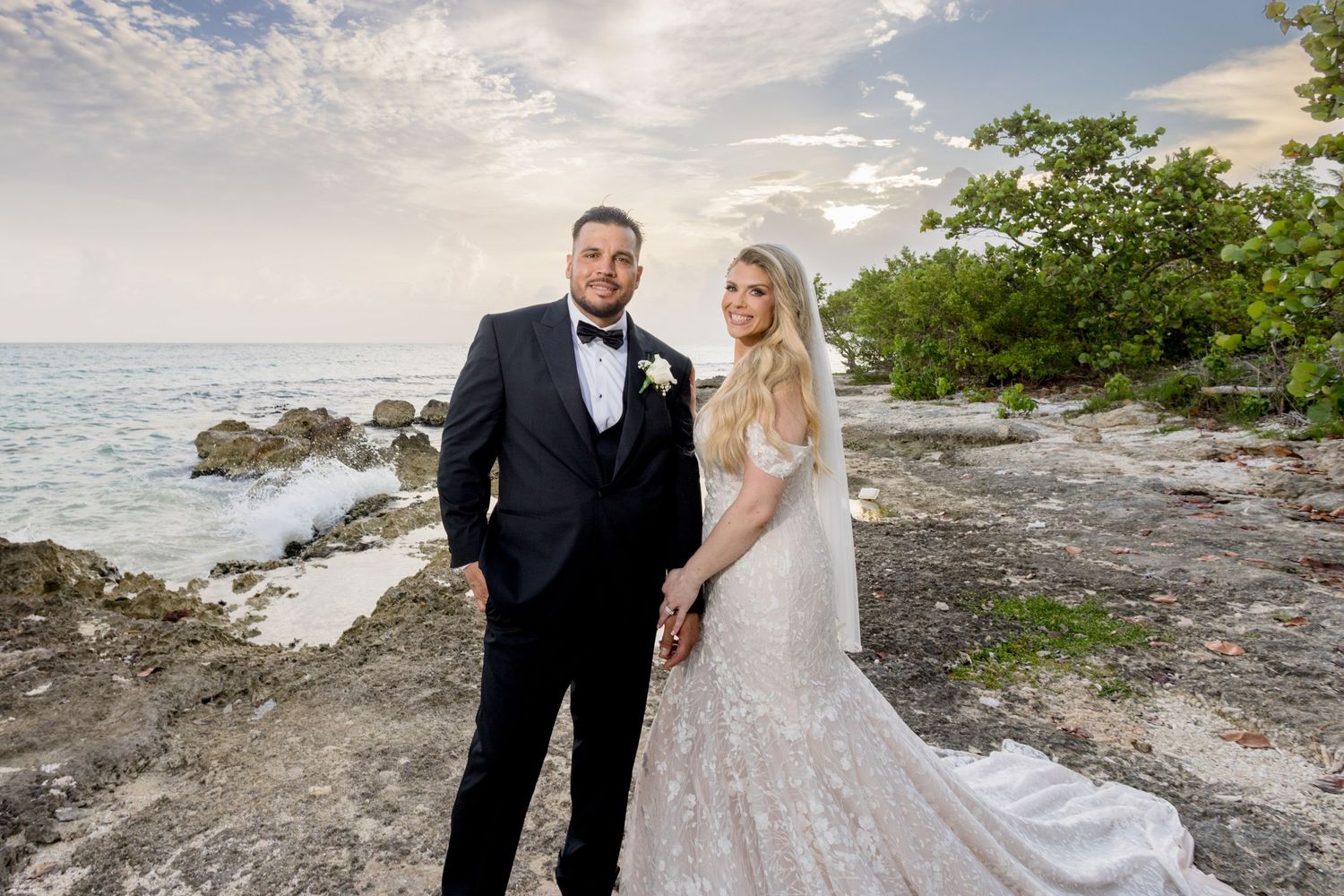 A couple in wedding attire poses for romantic photos along a rocky beach coastline at sunset. Hilton La romana