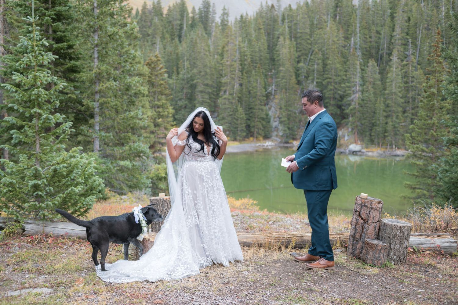 A bride holds her veil out of her face and looks back at her dog who stands near as her groom says vows to her near a mountain lake.
