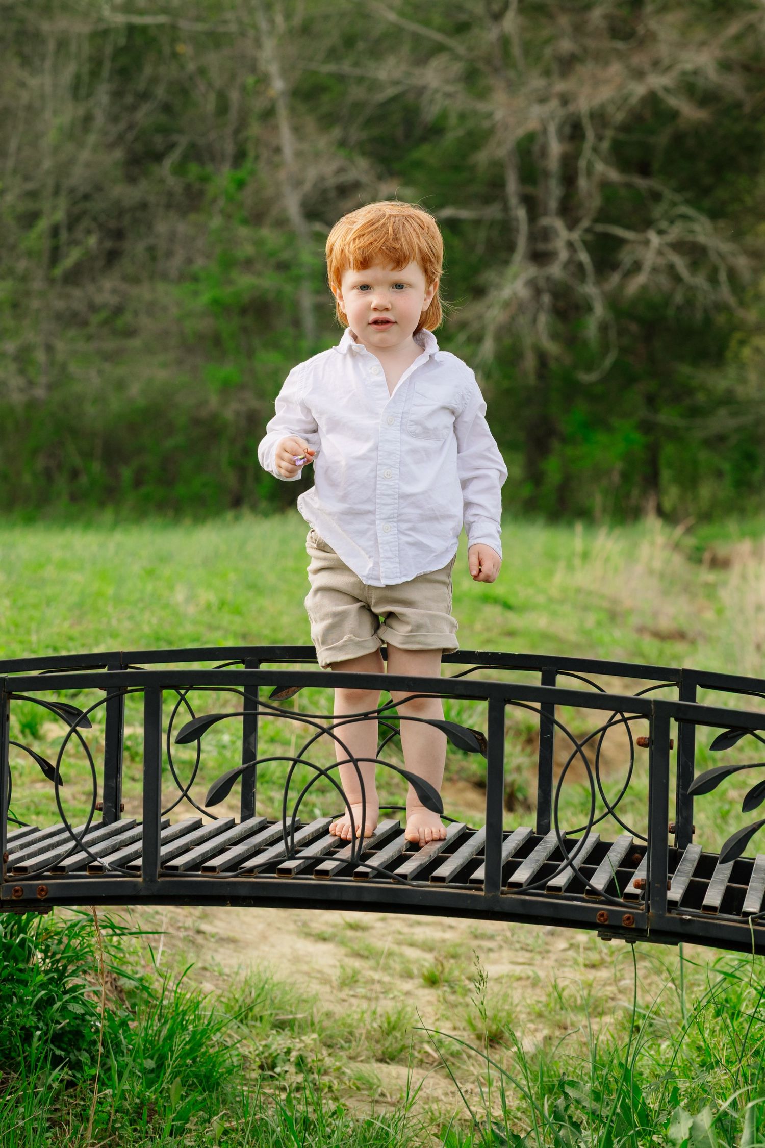 A child in a white shirt and khaki shorts walks along a decorative metal bridge in a lush garden setting.