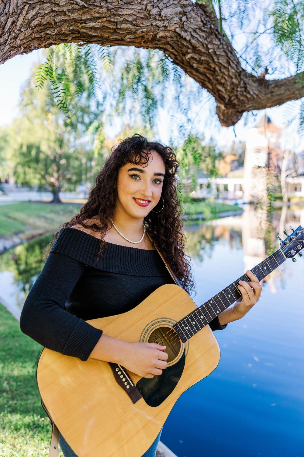 Woman in black sweater plays acoustic guitar outdoors by a lake on a sunny day.