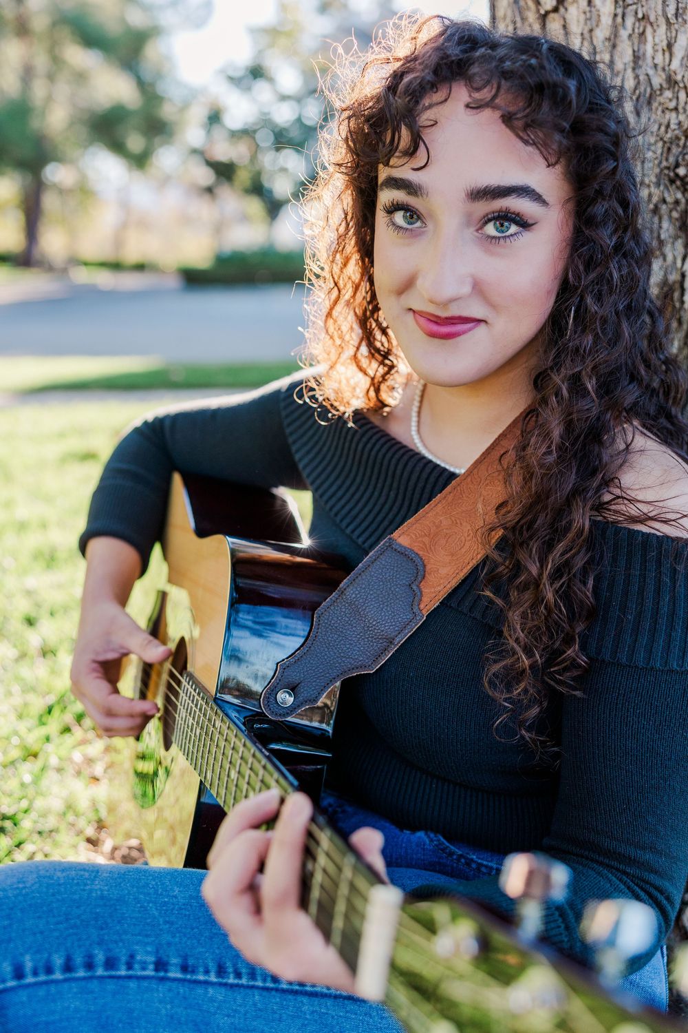 A musician in a black sweater plays acoustic guitar outdoors on a sunny day.