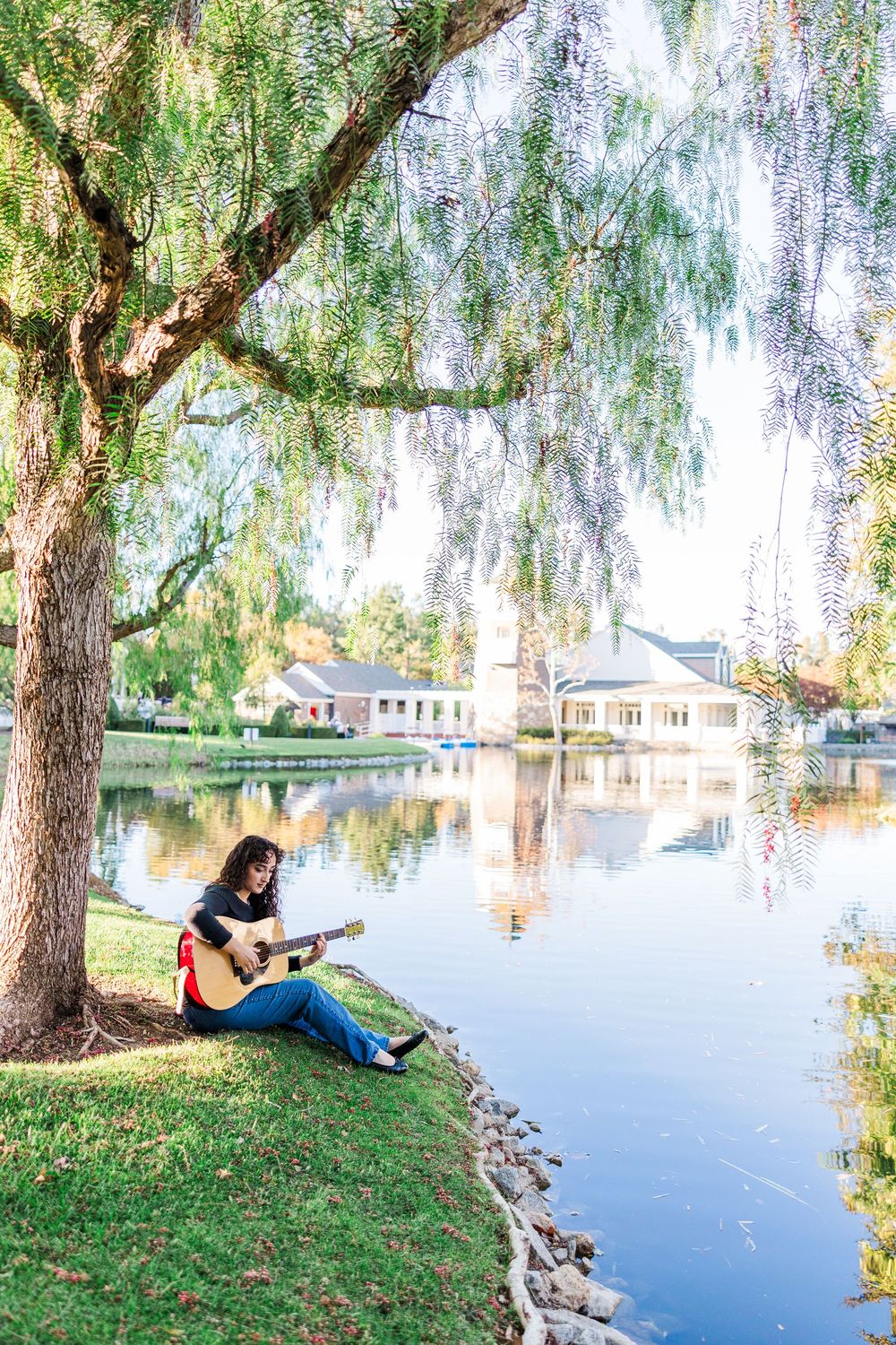 A person relaxes with a guitar by a tranquil lake under weeping willow trees on a sunny afternoon.