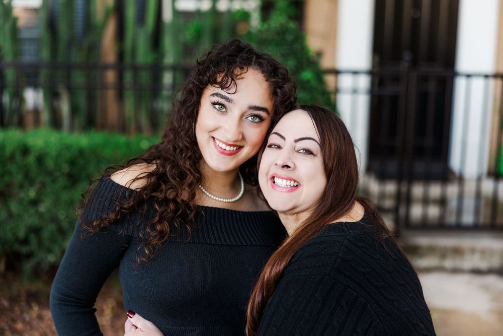 Two people with dark curly hair and straight hair embrace while smiling warmly at the camera outdoors.