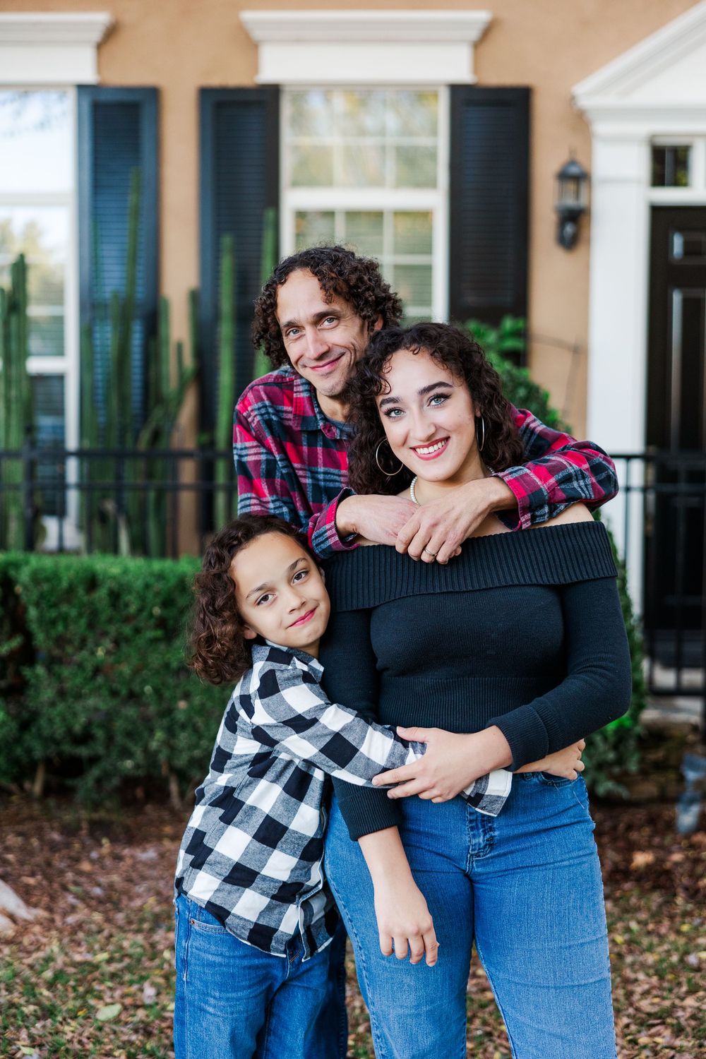 Three people in plaid shirts and jeans pose together affectionately outside a house.