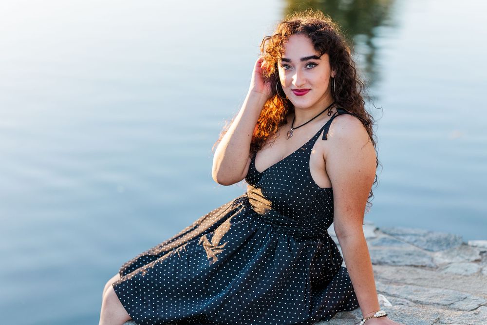 A woman in a polka dot dress sits by the water during golden hour with her hand near her face.
