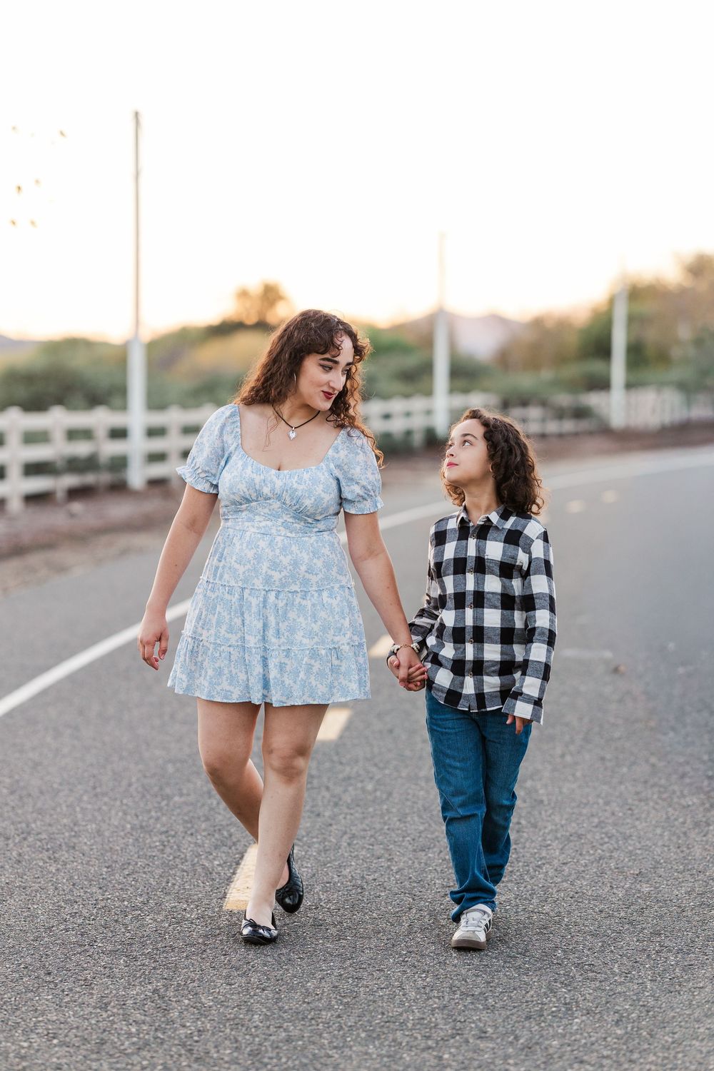 Two people walk together on asphalt road holding hands during golden hour, wearing blue and plaid outfits.