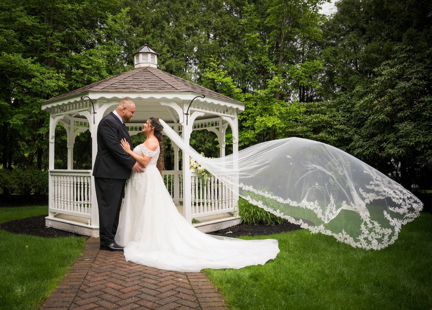 Wedding couple poses in front of white gazebo with flowing veil creating romantic garden atmosphere.