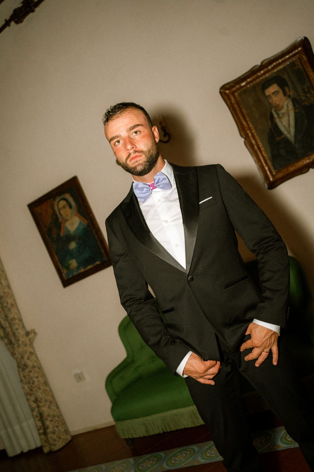 A person in a black suit and bow tie poses in different angles against a wall with vintage artwork in the background.
