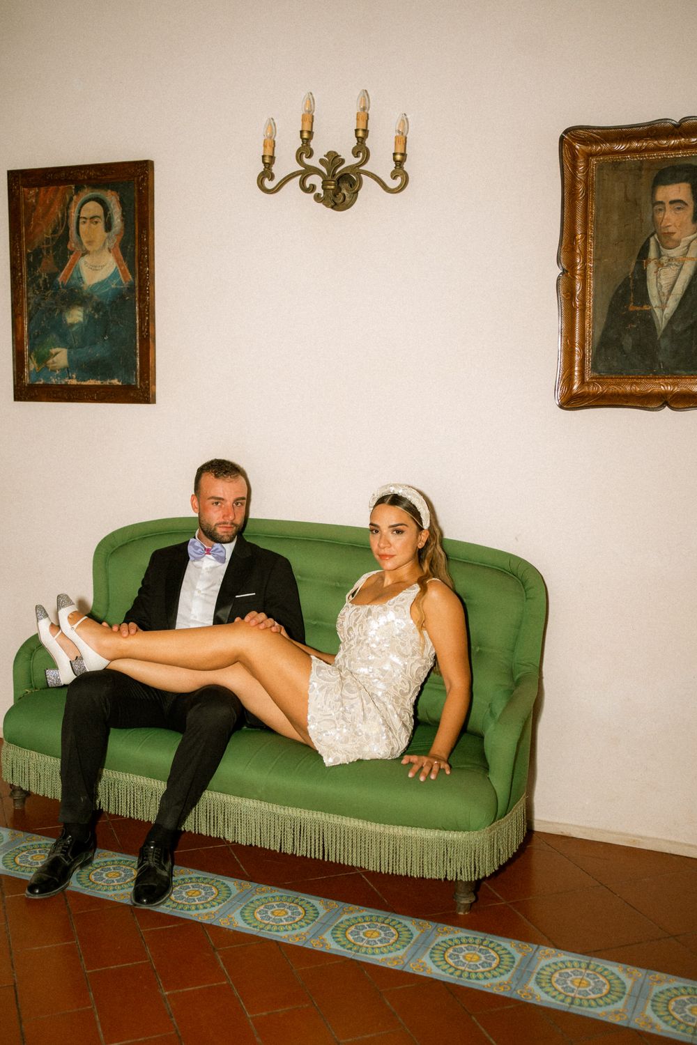 A couple in wedding attire relaxes on a vintage green couch in a room with portrait paintings on the wall.