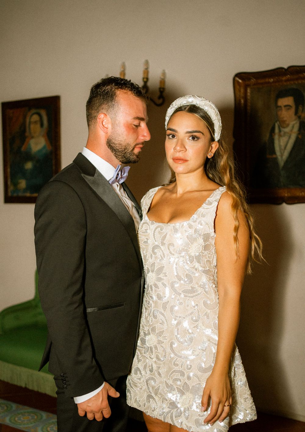 A romantic wedding couple in formal attire pose together against a neutral wall with vintage portraits in the background.