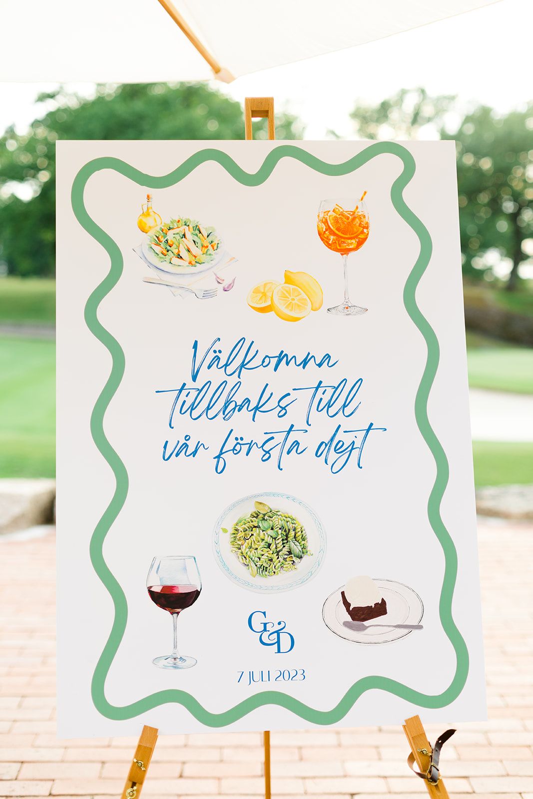 Watercolor wedding signs with blue calligraphy and green borders displayed on a brick patio with trees in background.
