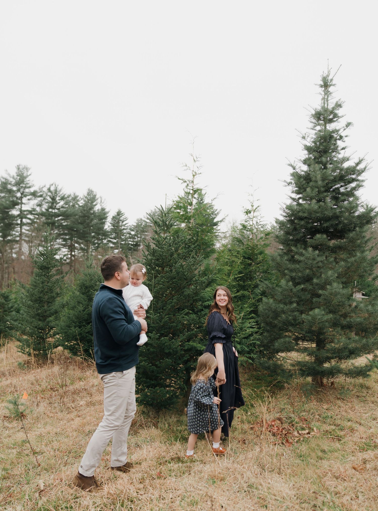 Family enjoys a winter outing at a Christmas tree farm surrounded by evergreen trees in a rustic setting.