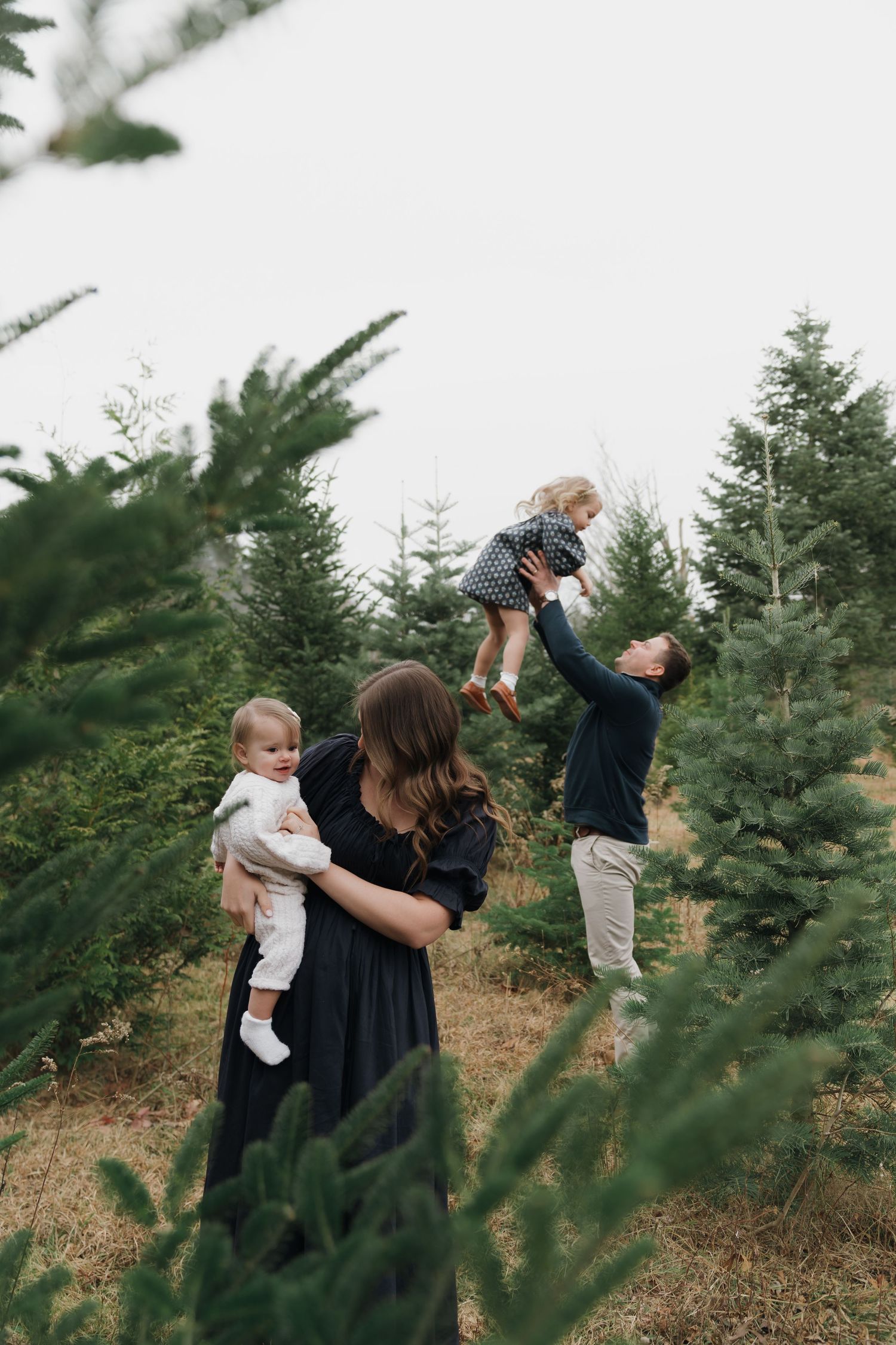 A family shares a tender moment together among evergreen trees at a Christmas tree farm.
