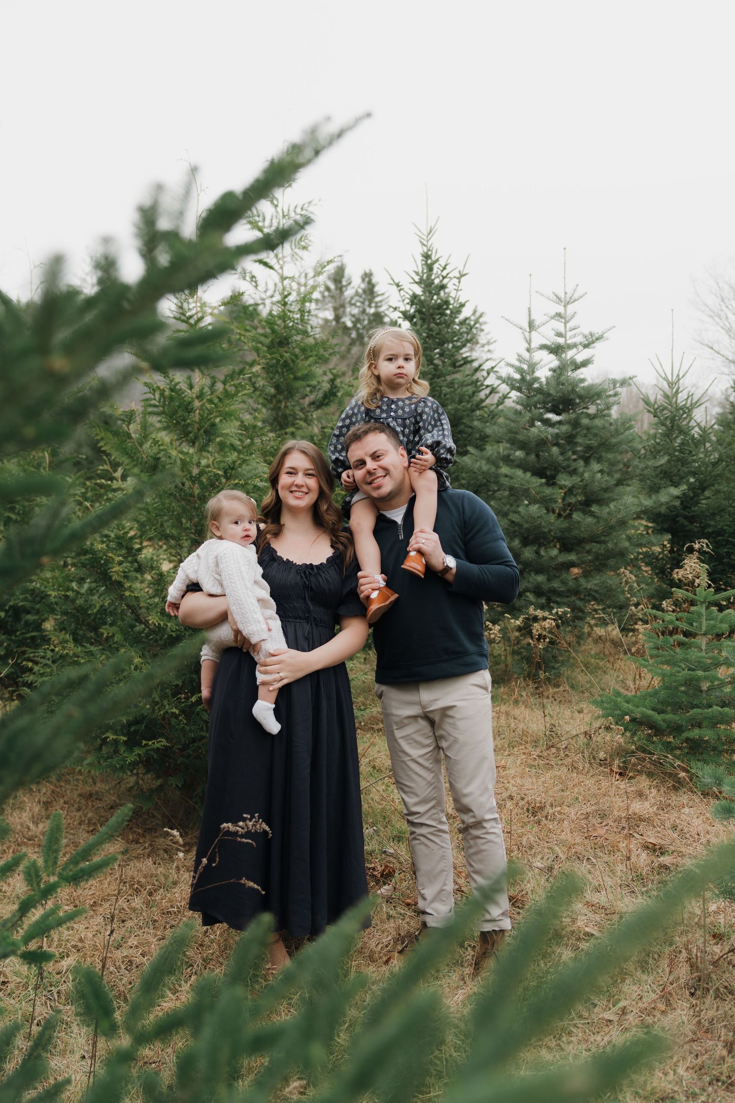A family poses together among evergreen trees at a Christmas tree farm on a cloudy day.