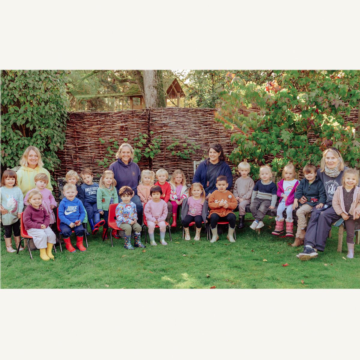 Preschool nursery photograph in an outdoor garden.  The teachers are sitting on chairs with the children around them.