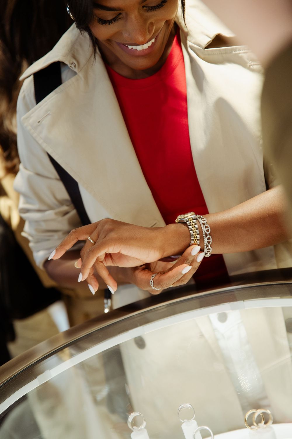 Smiling woman trying on rings at a jewelry store display case, wearing a red top and beige trench coat.