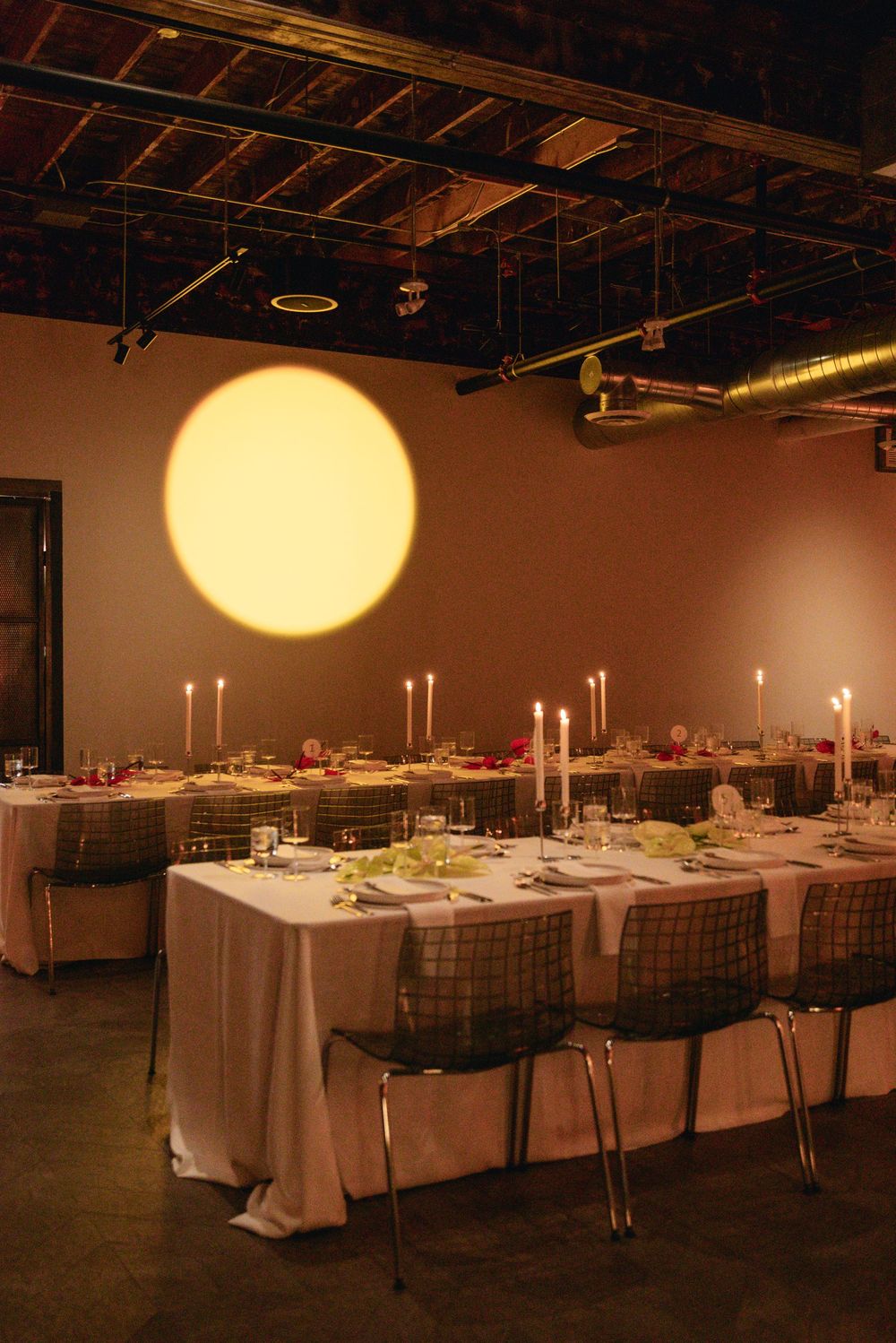 Elegant dining setup with long tables, white linens, and candlesticks under a warm circular light fixture.
