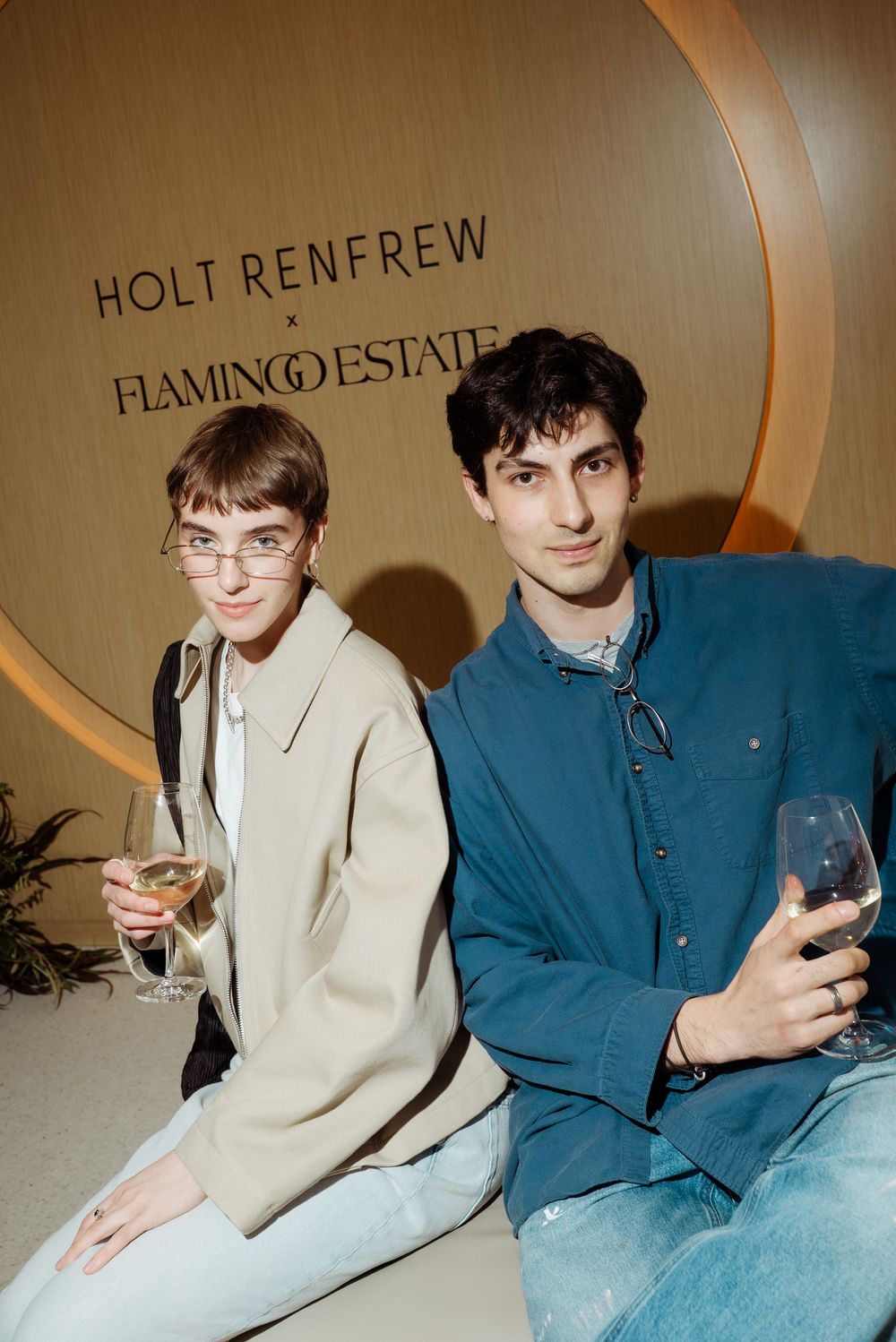 Two guests holding wine glasses at a Holt Renfrew x Flamingo Estate event, posing in front of a branded sign.