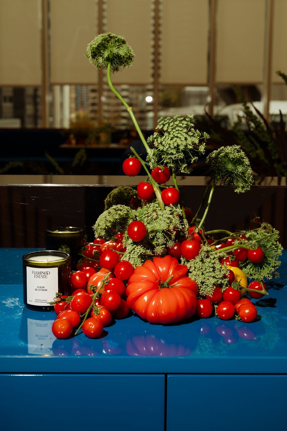 Fresh heirloom tomatoes and cherry tomatoes arranged with green herbs on a blue surface beside a candle.