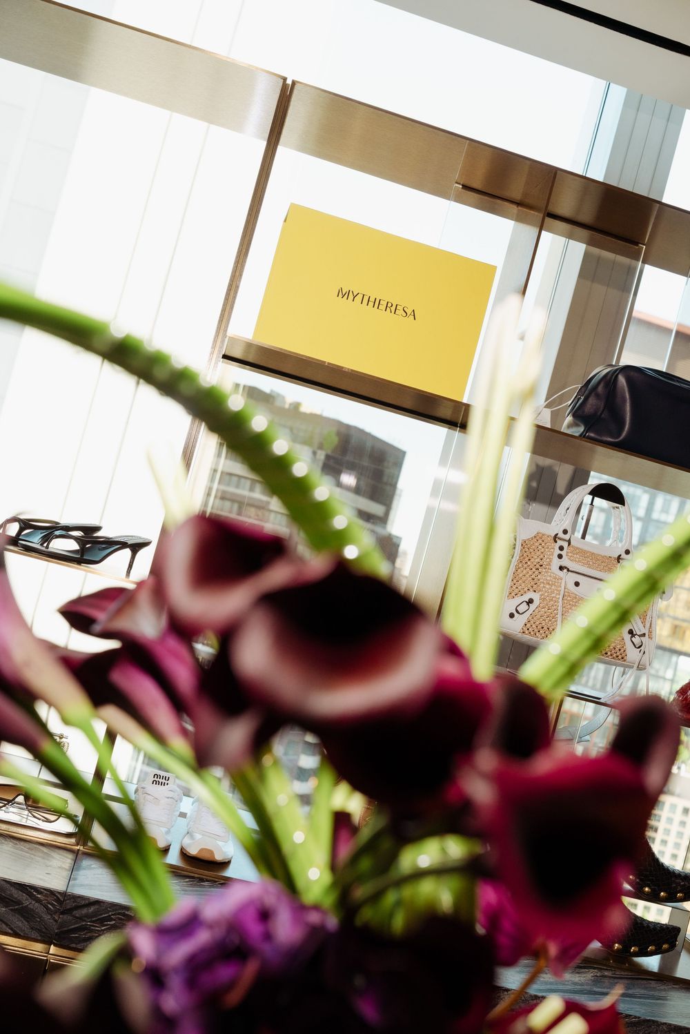 Mytheresa luxury boutique storefront with dark calla lilies in foreground and designer handbags displayed inside.