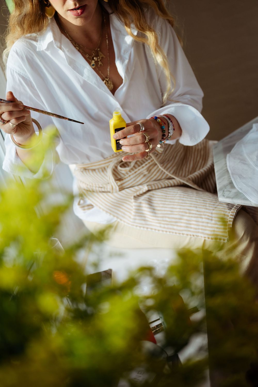 Woman in white shirt painting a striped bag with a brush, wearing gold jewelry and bracelets.