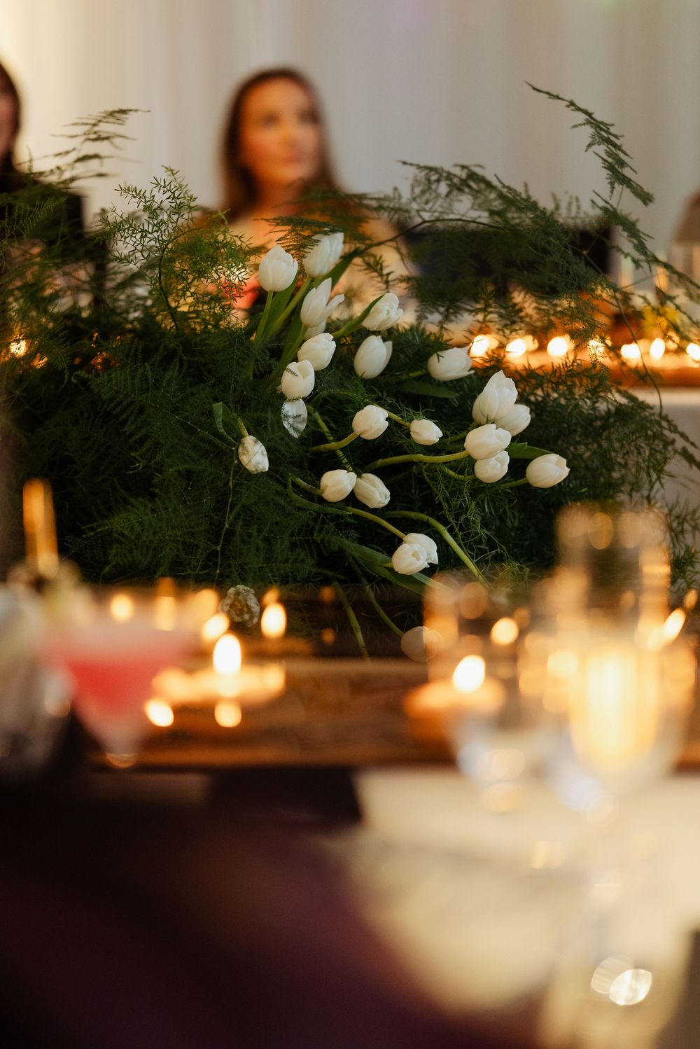 White tulips and greenery centerpiece with glowing candles at an elegant wedding reception dinner table.