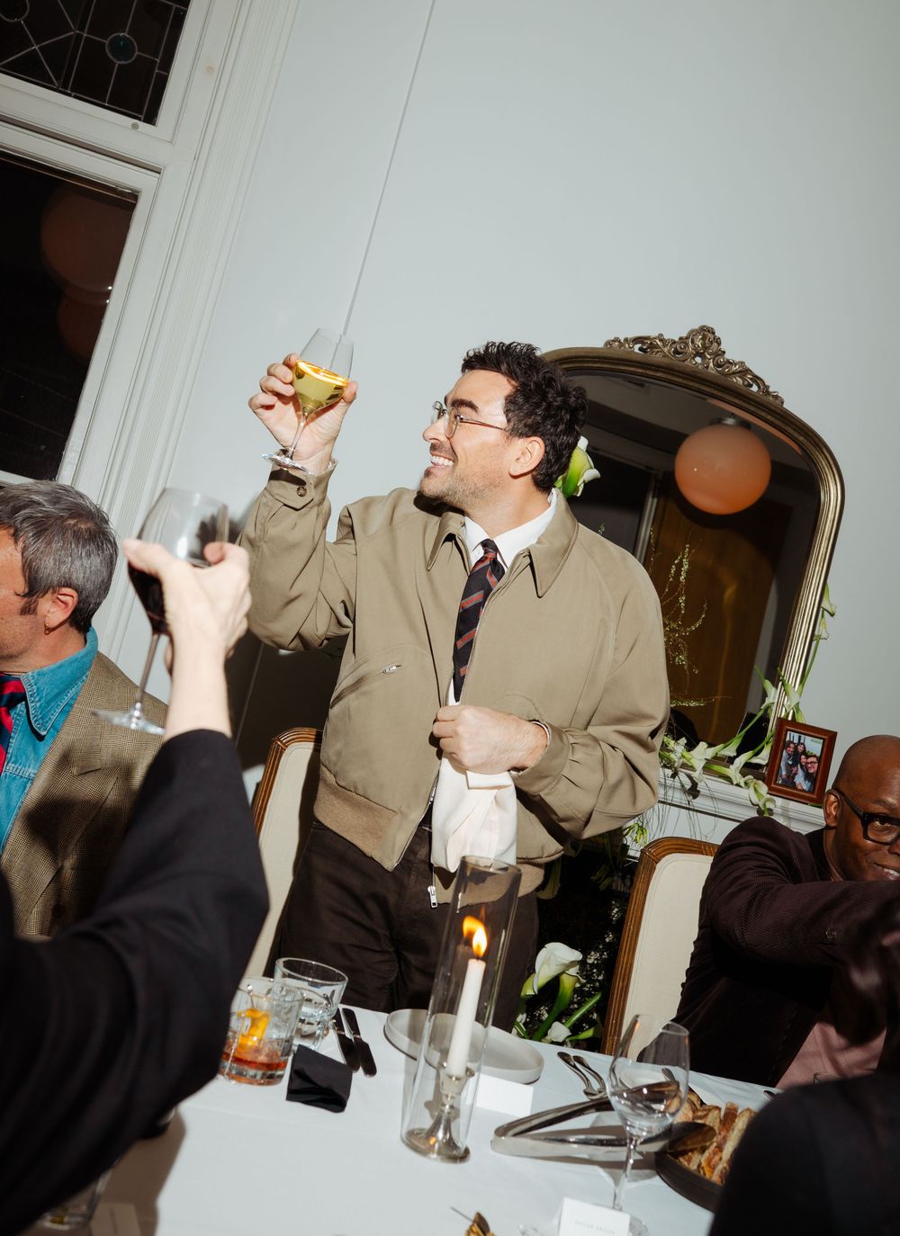 Man in beige jacket raises glass for a toast at an elegant dinner party with candlelit table and ornate mirror.