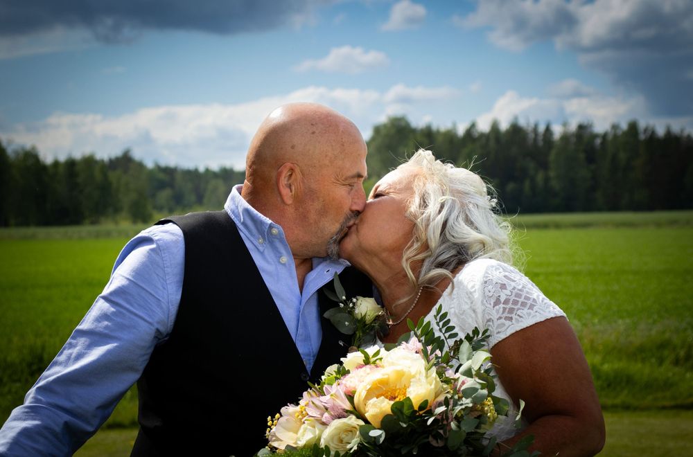 Brudepar kysser utendørs på sommerdag med blomsterbukett, romantisk bryllupsfoto av Prospectxmedia.